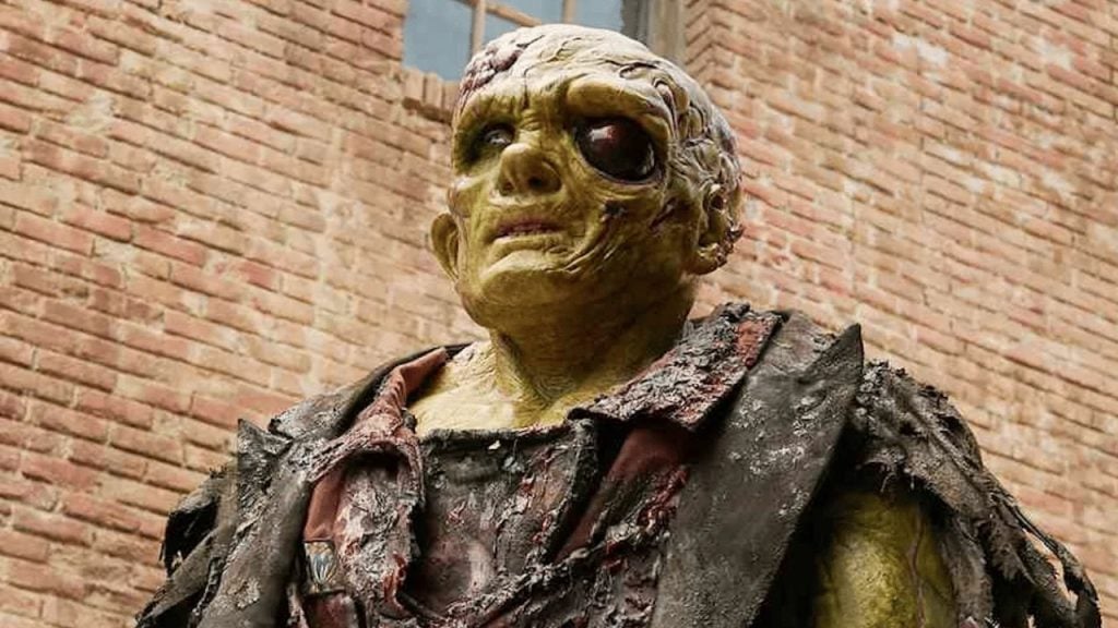 A person in detailed zombie makeup with yellowish skin, exposed teeth, and tattered clothing stands in front of a brick wall.
