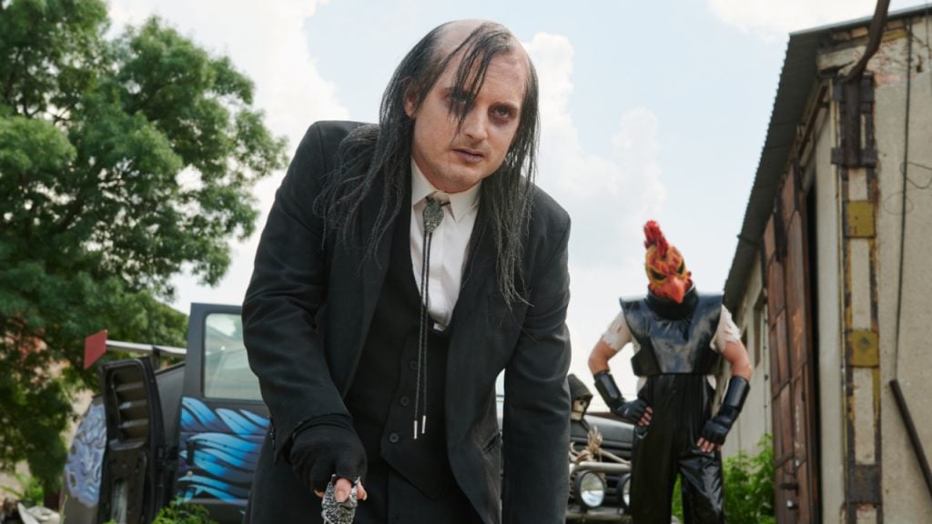A man in a black suit with long dark hair leans forward, holding a weapon, while another person in a flamboyant costume stands in the background near an outdoor building.