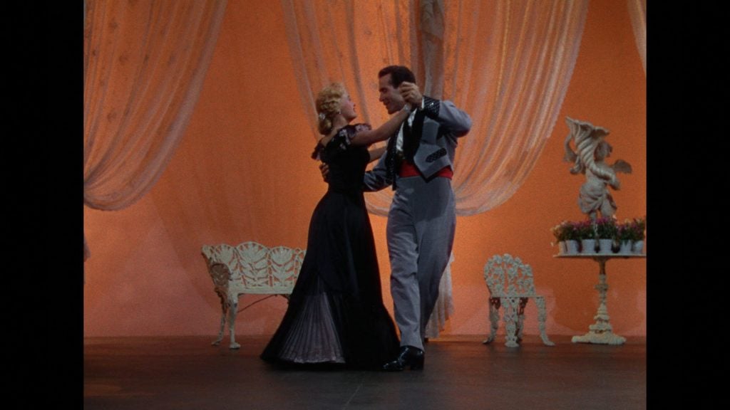 A man and a woman dressed formally are ballroom dancing on a stage with peach-colored drapes, white furniture, and a decorative statue in the background.