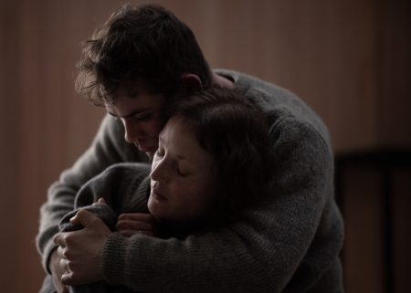 A man and a woman, both wearing sweaters, share a close, comforting embrace with closed eyes in a softly lit room.