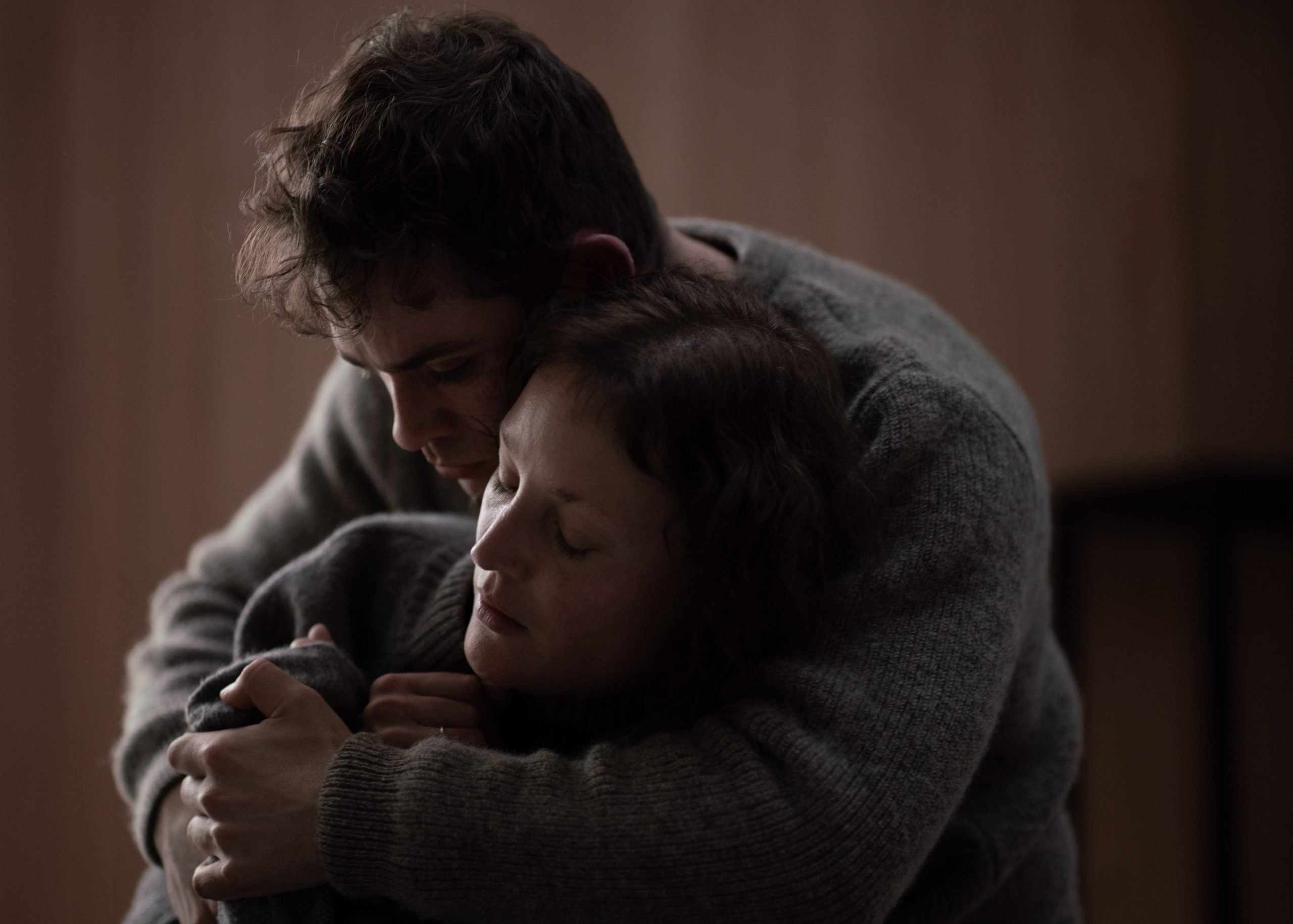 A man and a woman, both wearing sweaters, share a close, comforting embrace with closed eyes in a softly lit room.