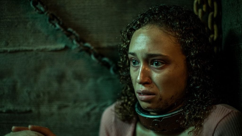 A woman with curly hair and a distressed expression sits against a wooden wall, wearing a metal collar attached to a heavy chain.