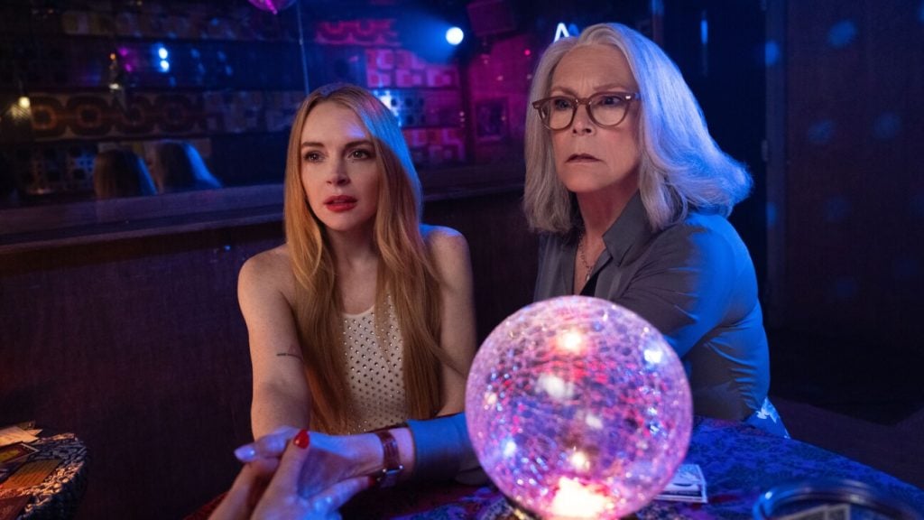Two women sit at a table in a dimly lit room, looking intently at something off-camera. A glowing crystal ball is on the table in front of them.