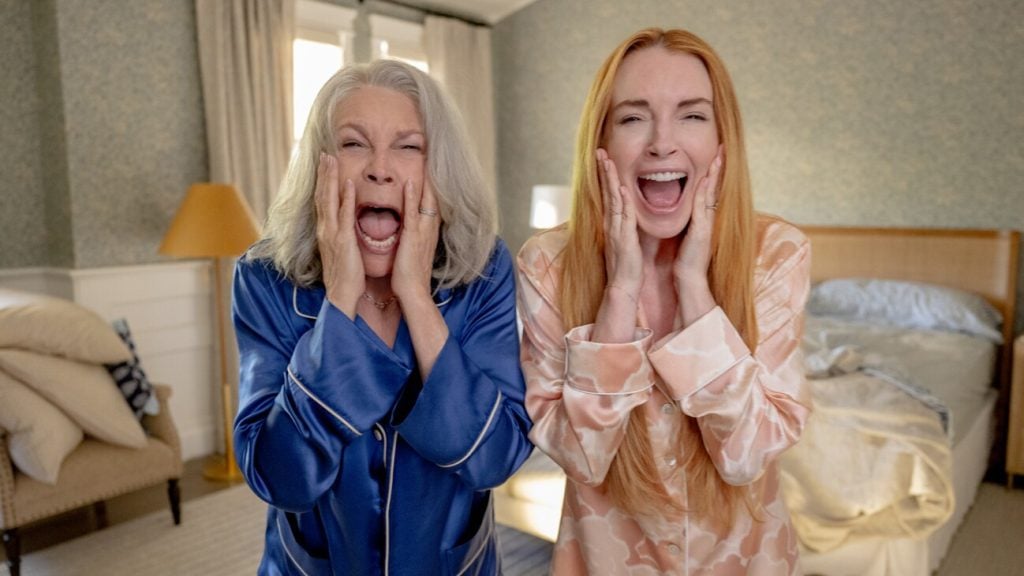 Two women in pajamas stand in a bedroom, facing the camera with their hands on their cheeks and mouths open as if screaming or expressing surprise.