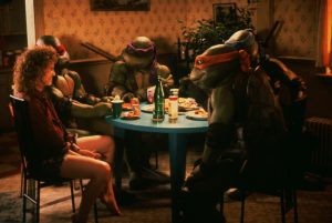 Four costumed Teenage Mutant Ninja Turtles sit around a table with a woman, eating pizza in a dimly lit, cluttered room.