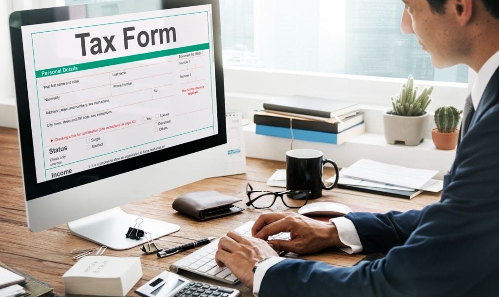A person in business attire fills out a tax form on a desktop computer at an office desk with various office supplies and documents.