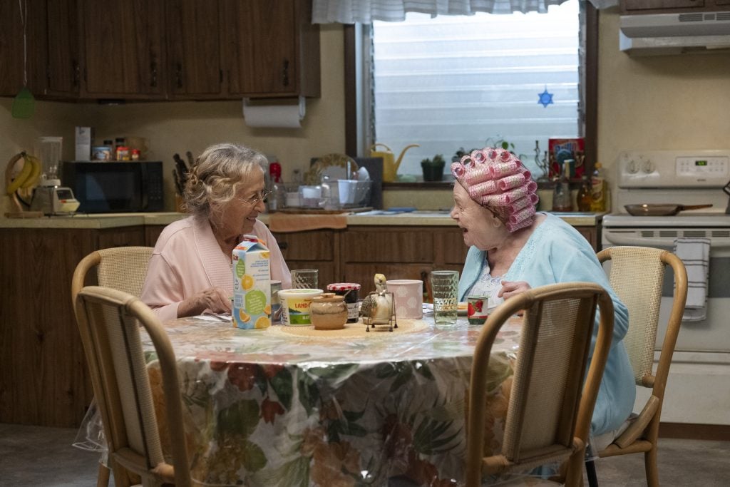 Two elderly women sit at a kitchen table eating breakfast and talking. One wears hair rollers. The table has food, drinks, and condiments. The kitchen is cozy and lived-in.