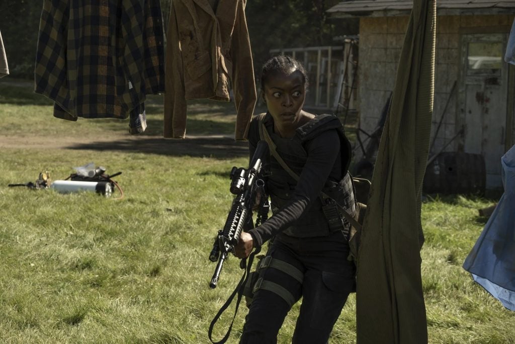 A person in tactical gear holds a rifle and crouches in a grassy yard near hanging clothes and a small building in daylight.