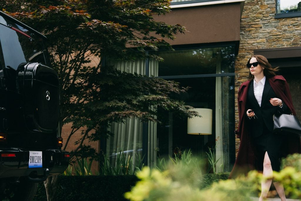 A woman in business attire and a maroon coat walks outside near a black SUV, holding a phone and bag, in front of a modern building with large windows.