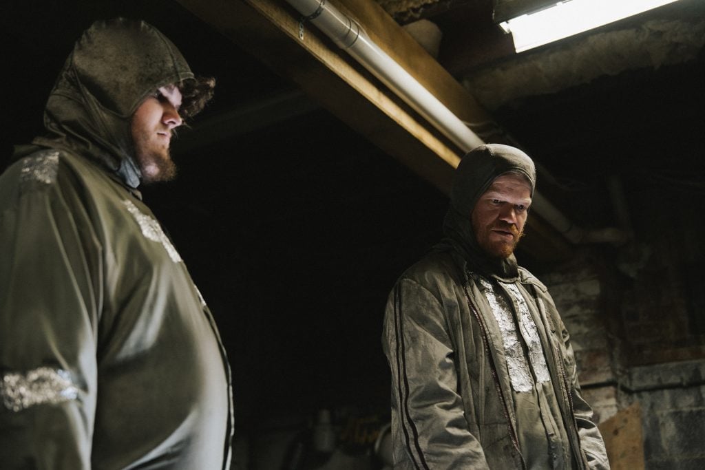 Two people wearing heavy, protective hooded suits stand indoors under artificial lighting, appearing focused on a task.