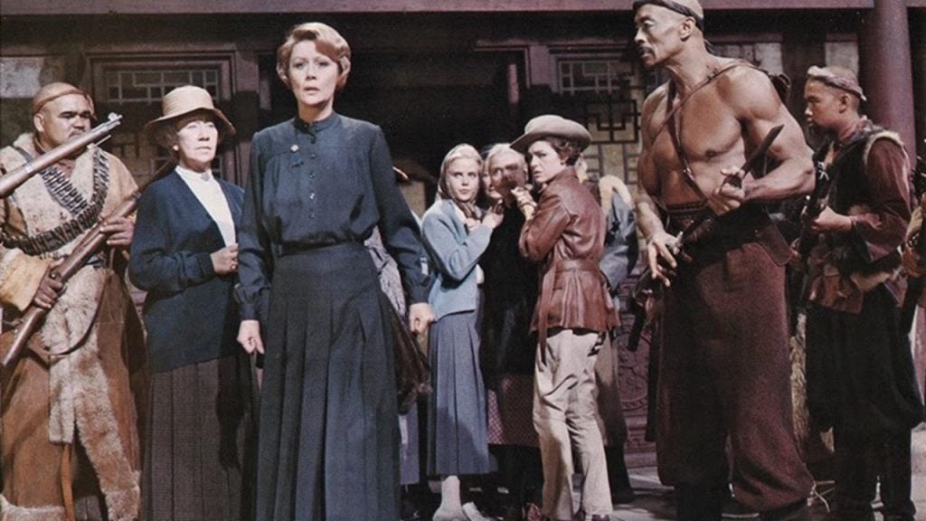 A group of people, including women in period clothing and a shirtless man with a spear, stand together in a tense scene, appearing alert and concerned.