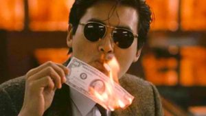 A man wearing sunglasses lights a cigarette using a burning U.S. hundred dollar bill.
