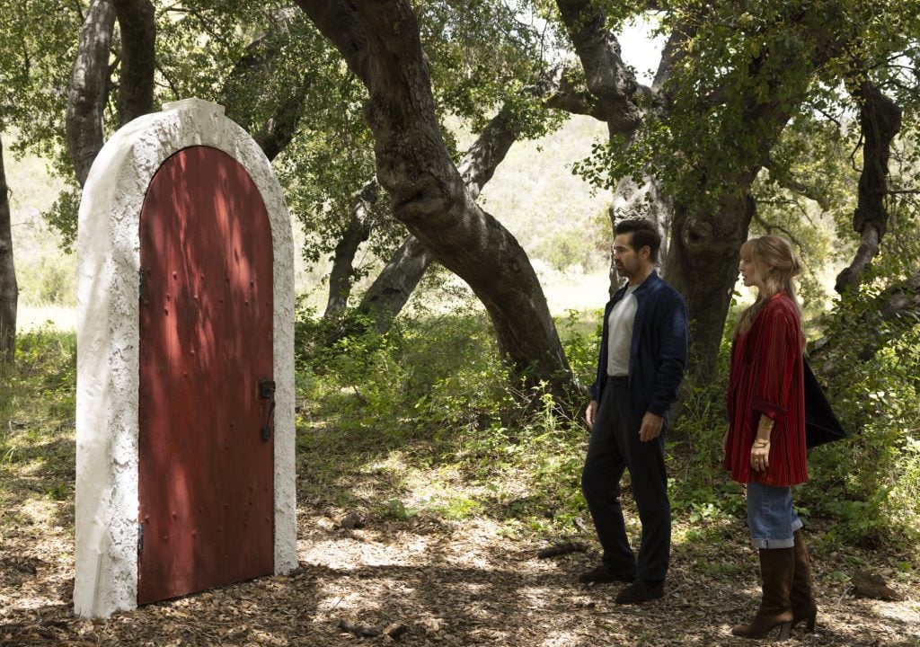 Two people stand in a wooded area, looking at a red arched door standing upright and freestanding among the trees.