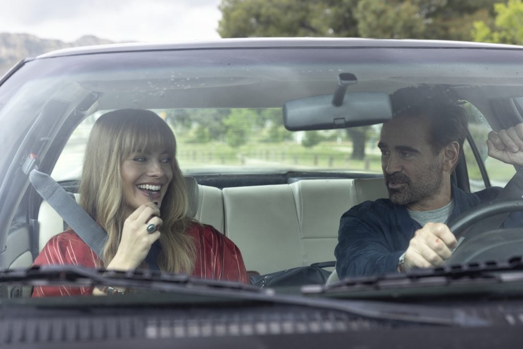 A woman in a red top and a man in a dark shirt sit in the front seats of a car; the woman is smiling and the man is driving.