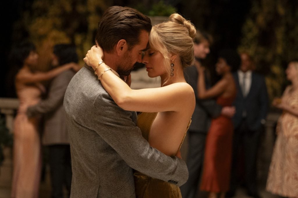A man and woman embrace closely while slow dancing at an elegant evening event, with other couples dancing in the background.