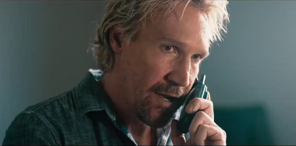 A man with blond hair and a beard holds a phone to his ear, speaking seriously indoors.