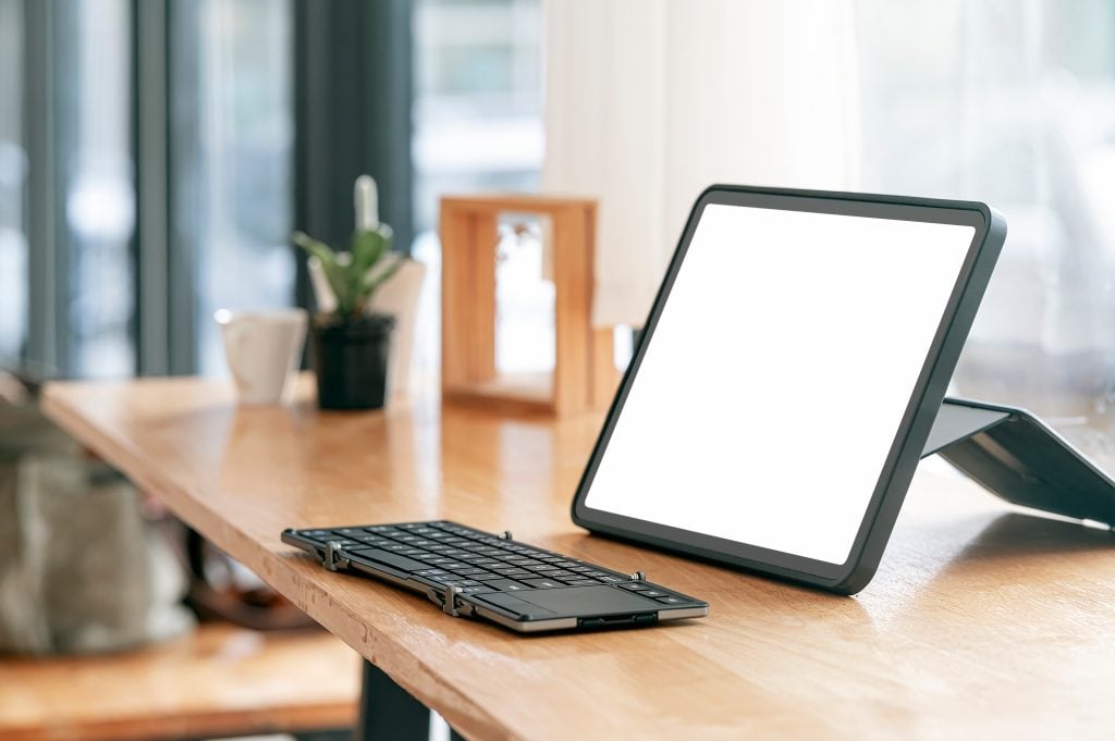 A tablet with a blank white screen sits on a wooden desk next to a detached keyboard, with a small plant and window in the background.