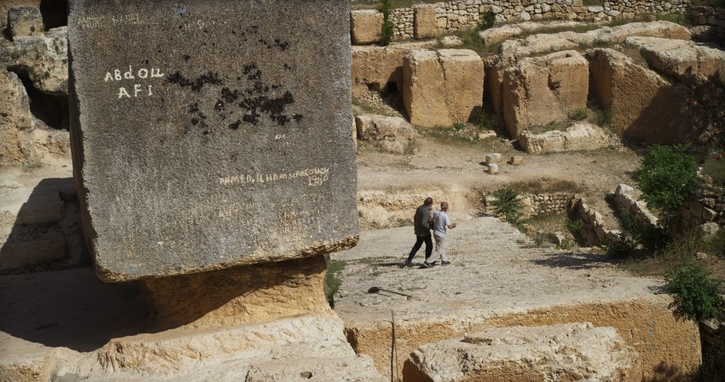 Two people walk near massive ancient stone blocks with graffiti on one, surrounded by weathered stone structures and sparse vegetation.