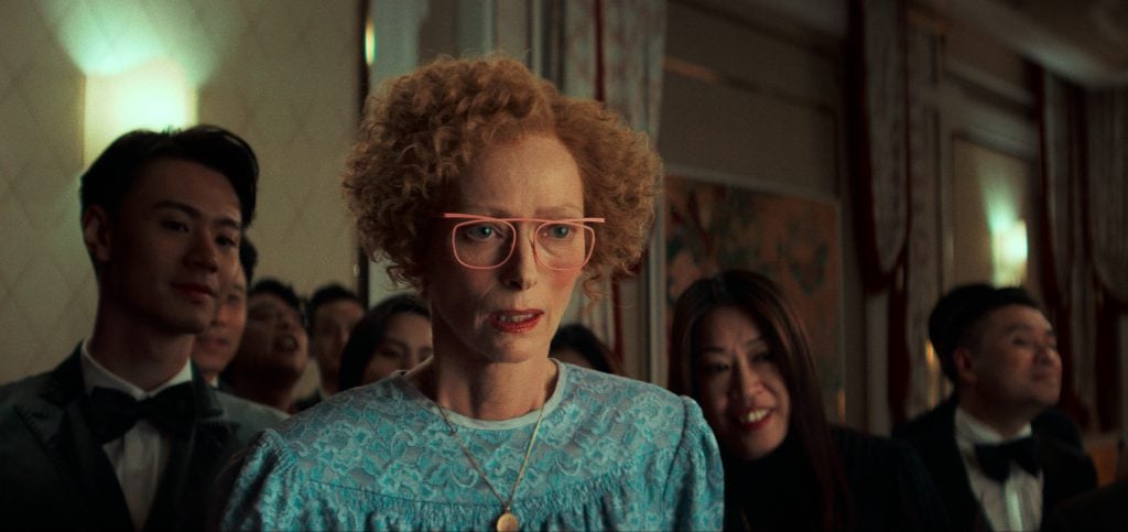 A woman with short curly hair and pink glasses wears a blue patterned dress, standing among a group of people in formal attire at an indoor event.