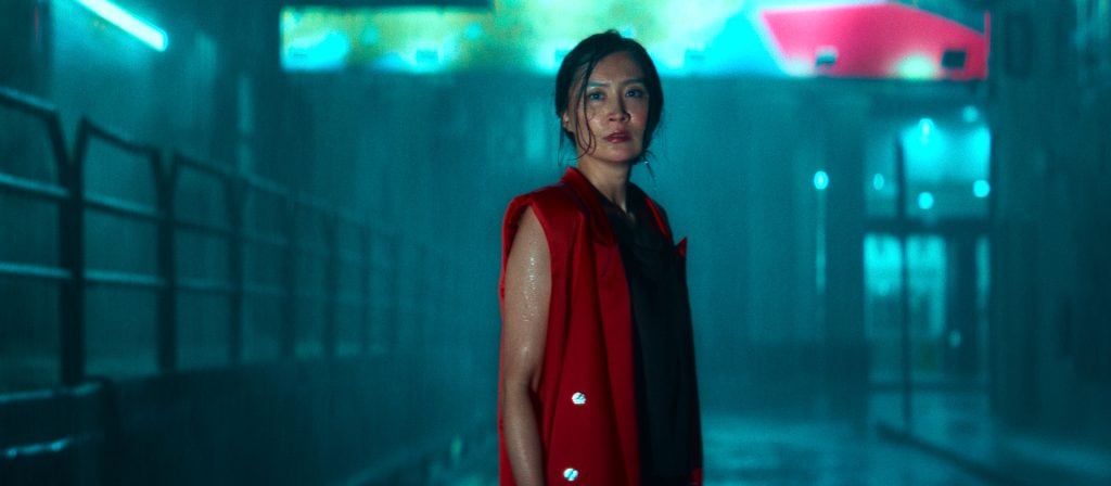 A woman in a red vest stands in a dimly lit, rainy urban alley at night, illuminated by blue and pink neon lights.