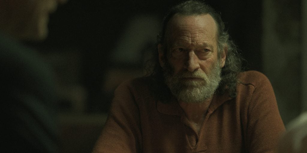 A man with long hair and a gray beard wearing a rust-colored shirt sits at a table, looking to his left with a serious expression.