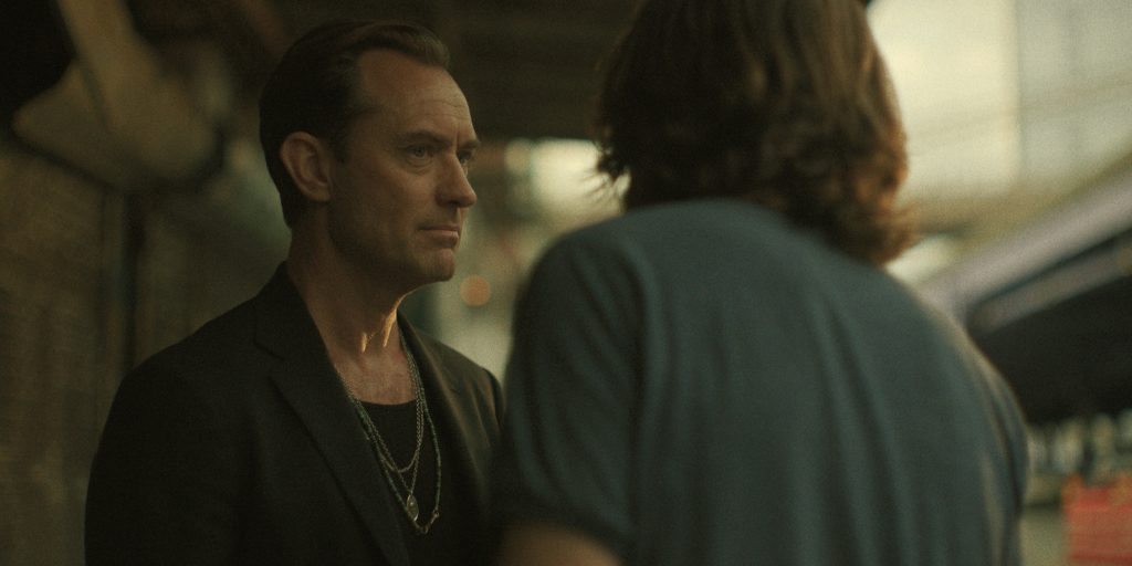 Two men stand facing each other outside, one in a dark blazer with necklaces and the other in a casual t-shirt, having a serious conversation.