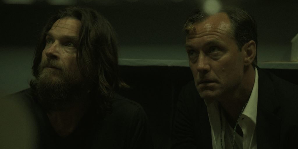 Two men sit close together in a dimly lit room, looking slightly upward with serious expressions.