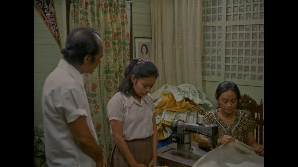 Three people are in a room; a woman sews fabric on a machine, a girl stands beside her with hands clasped, and a man stands nearby. Fabric and curtains are in the background.