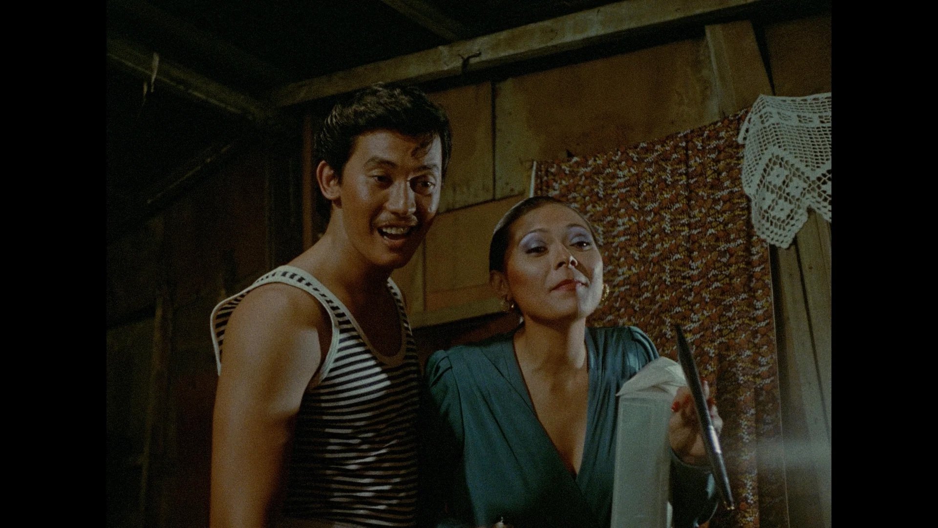 A man in a striped tank top and a woman in a green dress stand together indoors, both smiling. The woman holds a newspaper; the background shows patterned fabric and a crocheted doily.