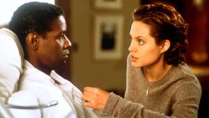 A woman in a sweater leans in to speak to a man lying in a hospital bed, looking at him with concern.