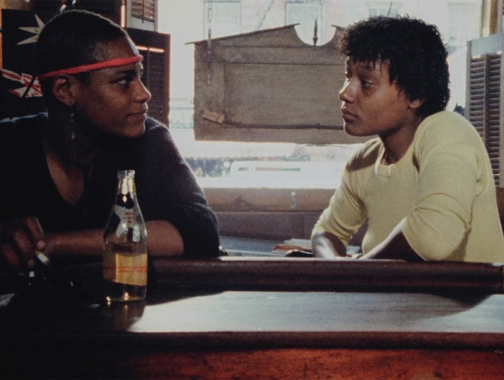 Two people sit at a wooden bar, facing each other and talking. A partially full glass bottle is on the bar in front of them, with sunlight coming through the window behind.