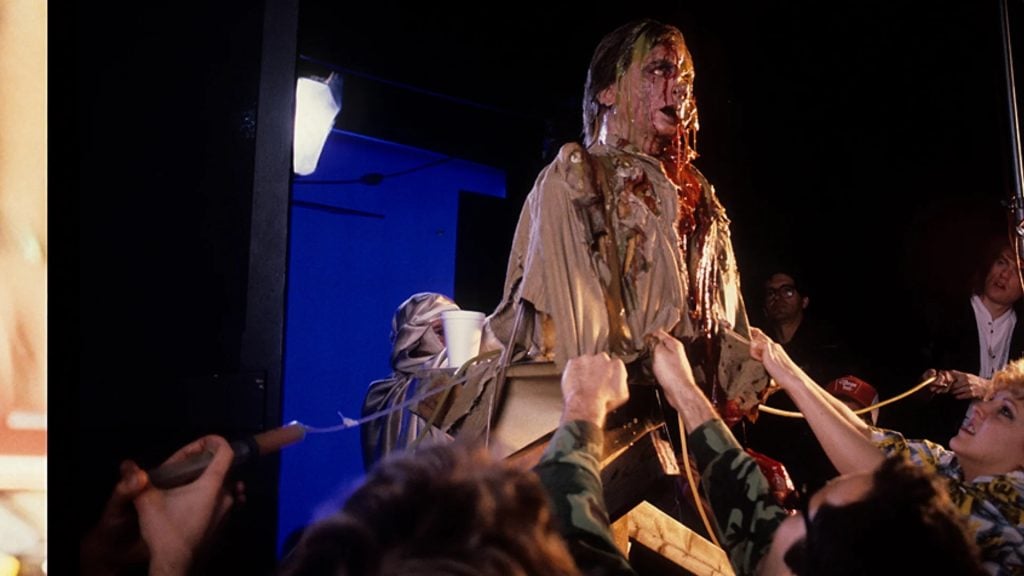 Several crew members work on a film set with a practical effect showing a person in a tan coat covered in fake blood and injuries, surrounded by filming equipment and blue screens.