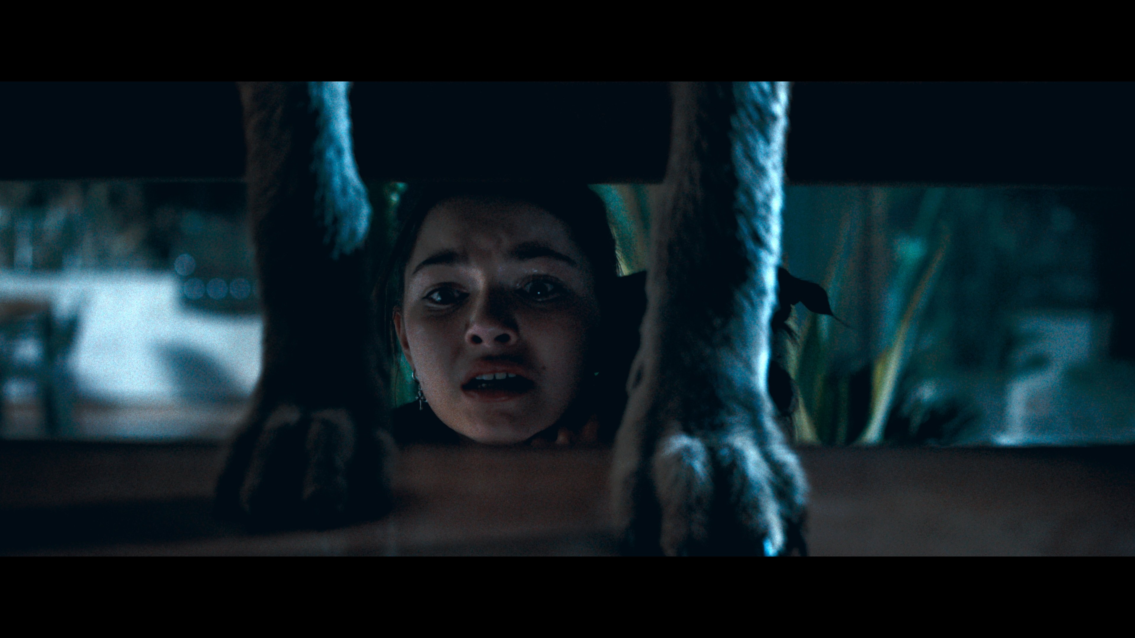 A frightened girl hides under a table as large animal paws stand on top of it, with her face illuminated by dim blue light.