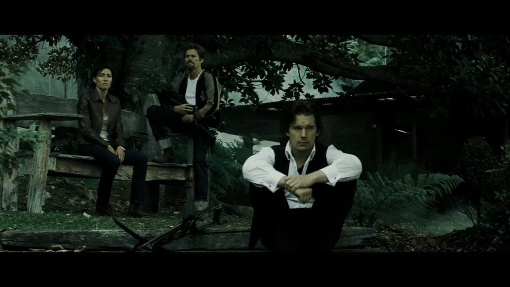 Three people sit and crouch outdoors near a large tree and a rustic wooden structure, with a somber atmosphere and subdued lighting.