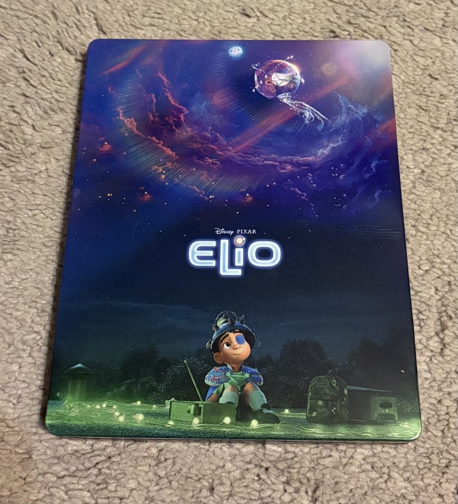 A Disney Pixar "Elio" poster showing a boy sitting on grass at night, looking up at a purple and blue sky with a spaceship and a large swirling nebula above him.