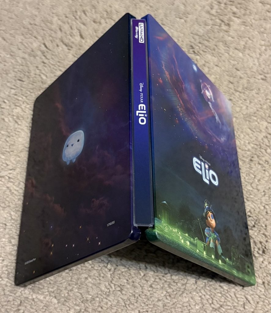 A Disney Pixar "Elio" steelbook case is standing upright and open on a carpeted floor, displaying both the front and back cover artwork.