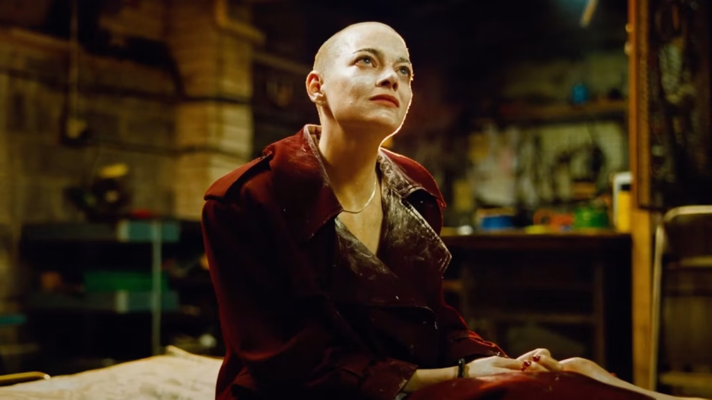 A bald person wearing a red coat sits on a bed in a dimly lit, cluttered room, looking upward with a serious expression.