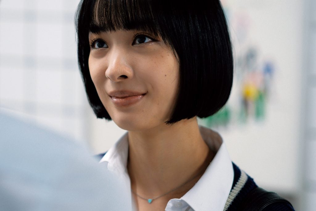 A woman with a short black bob haircut, wearing a white collared shirt and a necklace, looks slightly upward with a subtle smile in a brightly lit room.