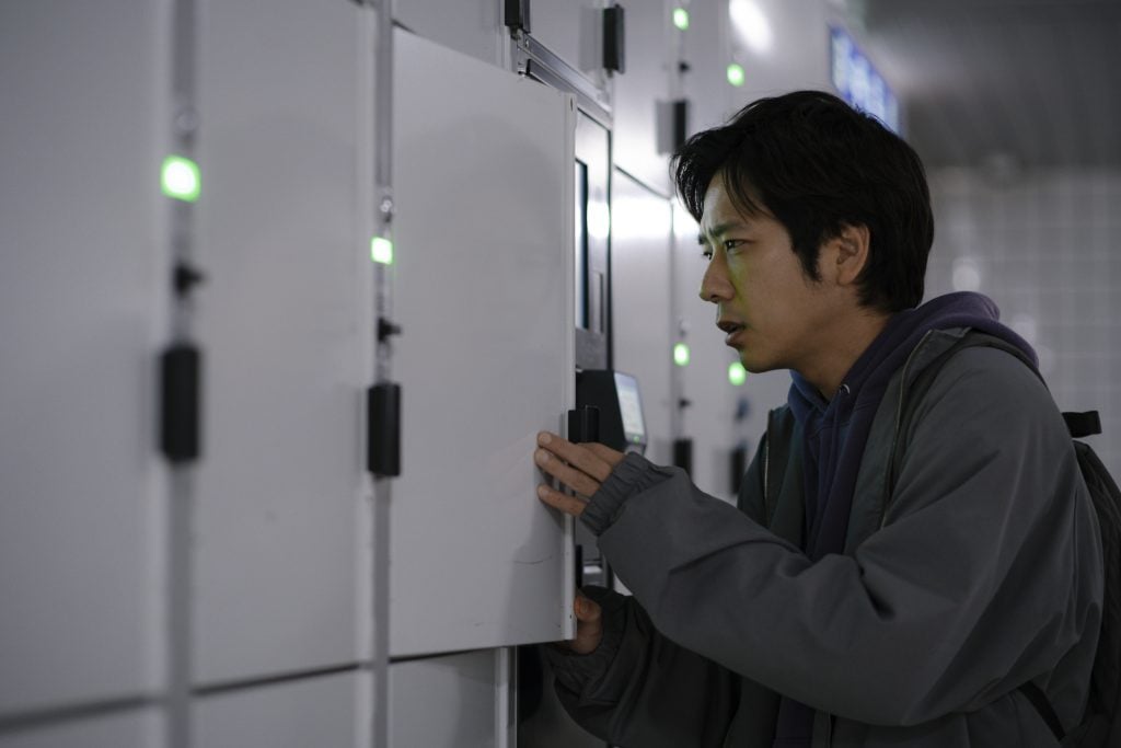 A man stands in front of a row of lockers, opening one as he looks inside with a focused expression.