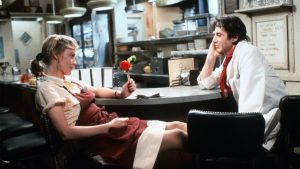 A waitress in uniform sits at a diner counter holding a flower, while a man in a white coat sits across from her, both appearing engaged in conversation.
