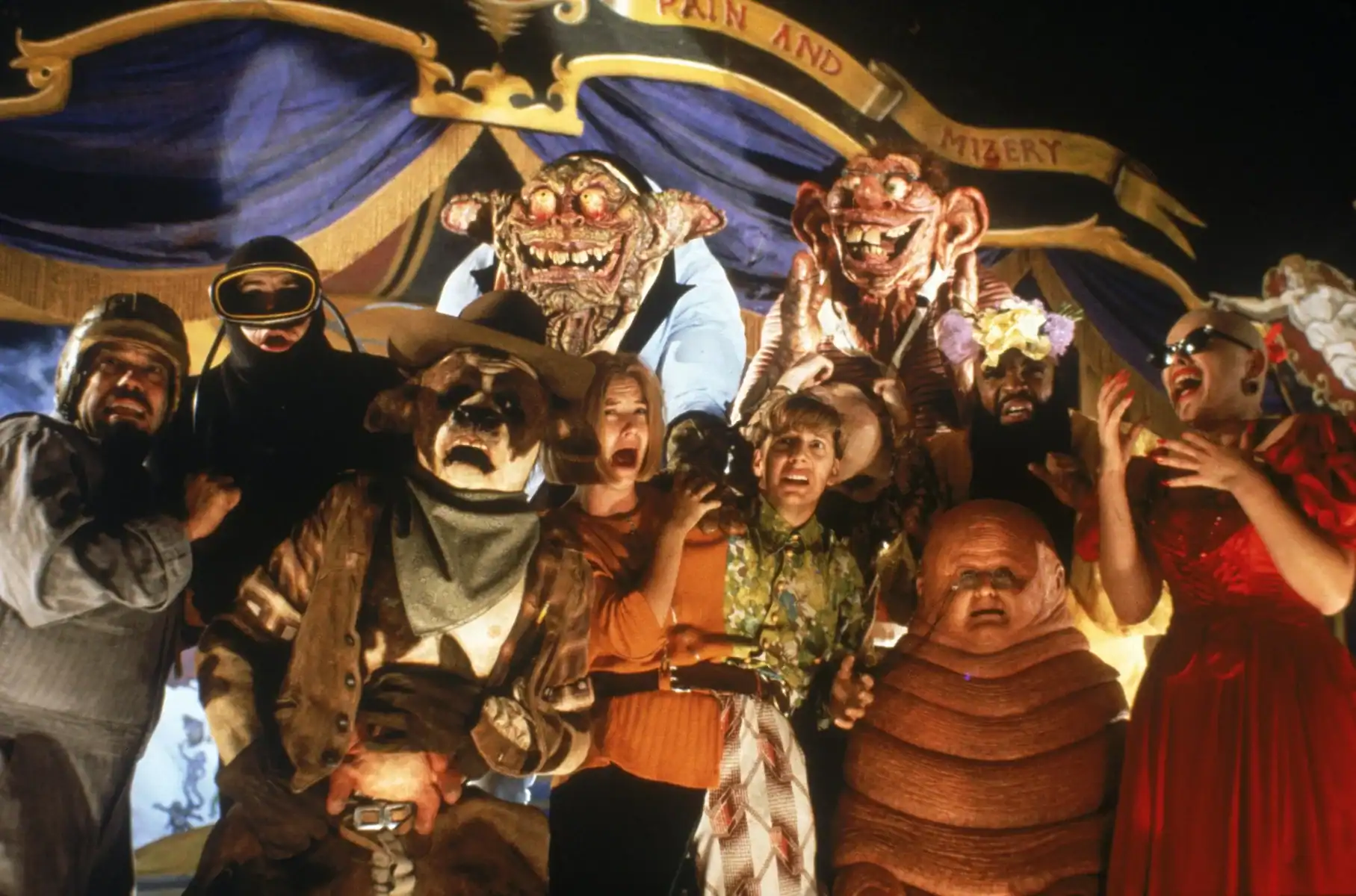 A group of people and grotesque puppet creatures pose together onstage in front of a circus-themed backdrop reading "Pain and Mizery.