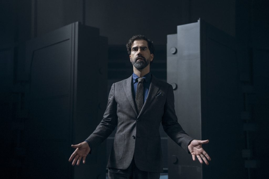 A man in a suit stands with arms slightly outstretched in front of large open metal doors, in a dimly lit setting.
