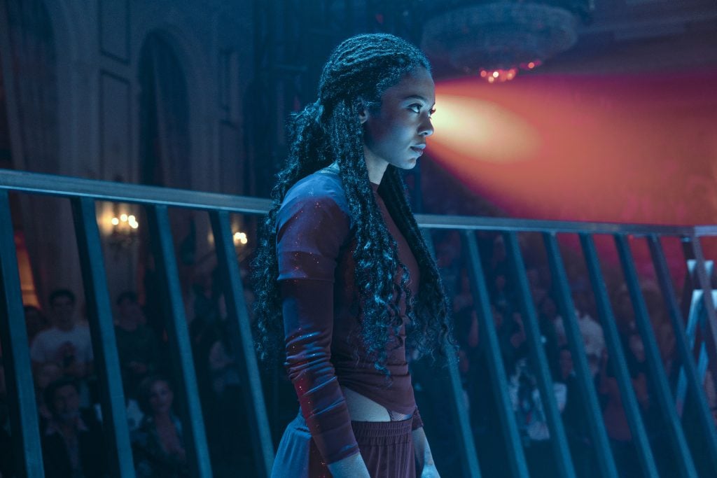 A woman with long braided hair stands in profile under dramatic red and blue lighting, with a crowd and a metal fence visible in the background.