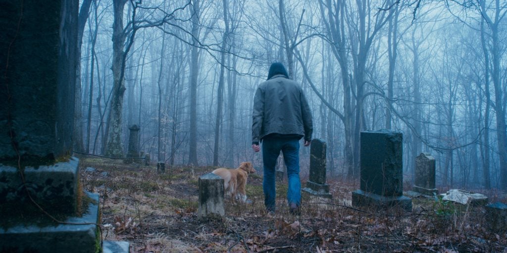 A person in a hooded jacket walks a dog through an old, foggy graveyard surrounded by bare trees.