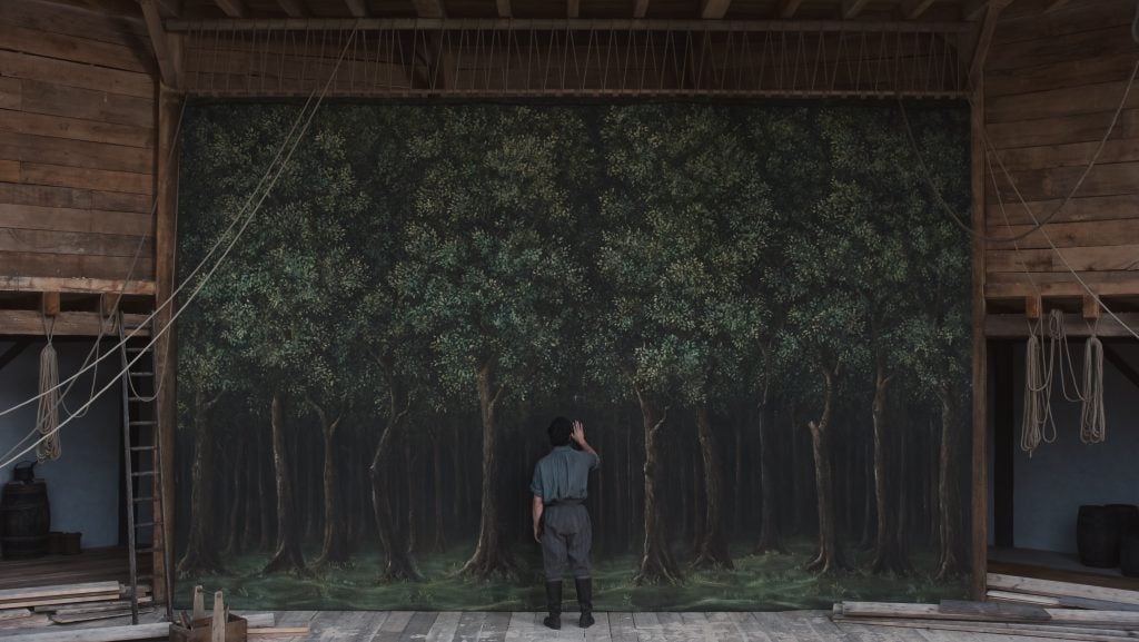 A person in period clothing stands on a wooden stage, facing and touching a large painted backdrop of a dense forest.