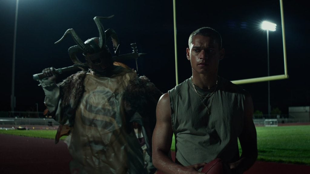 A young man holding a football stands on a field at night, with a costumed figure in a horned mask and fur behind him under stadium lights.