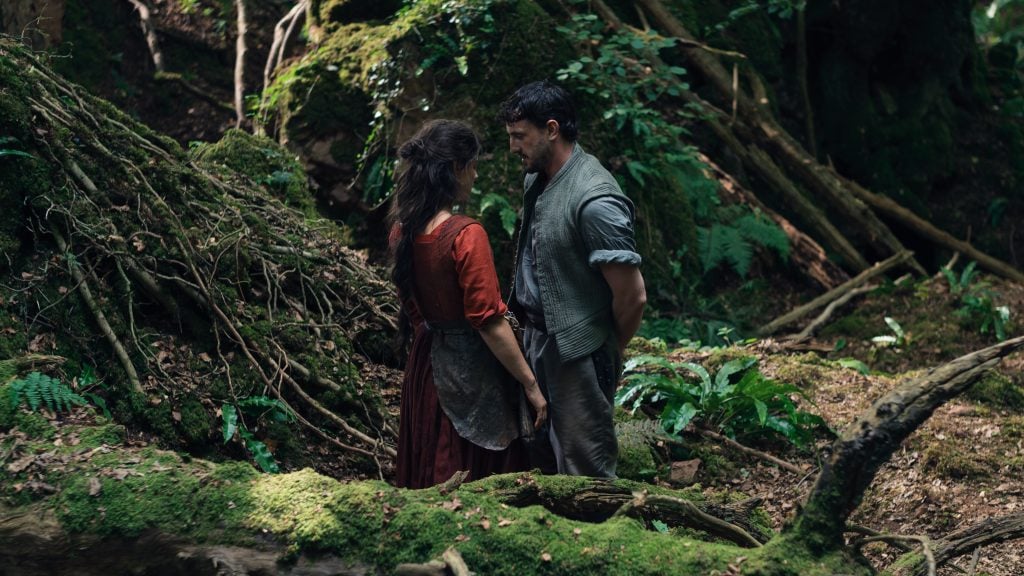 A man and woman stand facing each other in a dense, mossy forest surrounded by trees and greenery.