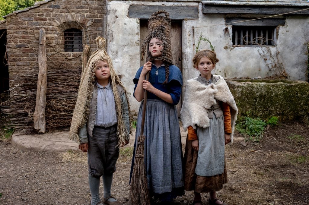 Three children in old-fashioned, rustic clothing stand outside a stone and plaster building, with two wearing woven baskets on their heads and one draped in a wool covering.