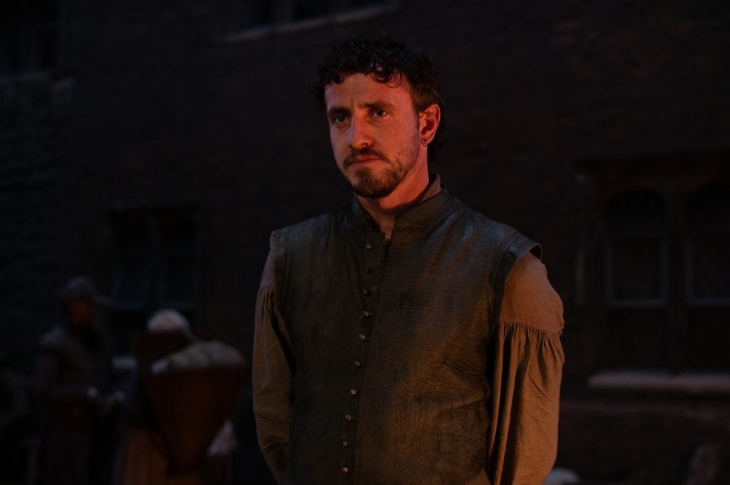 A man in historical clothing stands indoors with a serious expression; two blurred figures are seen in the dimly lit background.