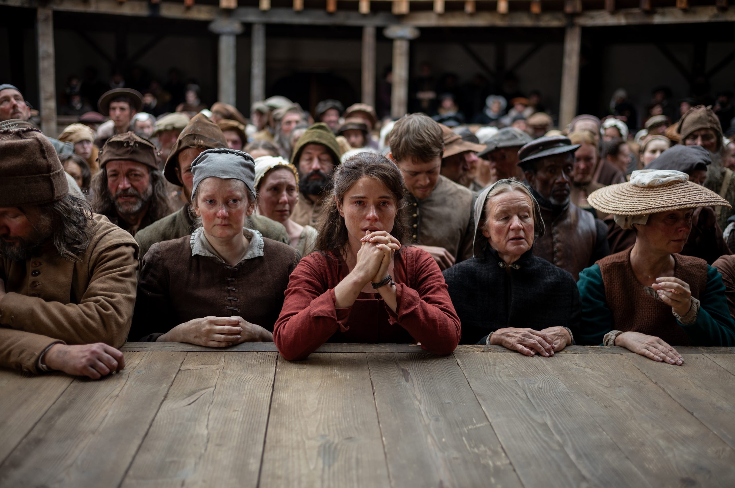 A large group of people in historical clothing stand closely together, watching intently from behind a wooden barrier in what appears to be a period setting.